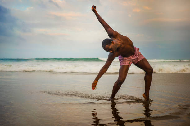 dramatic contemporary dance choreographer doing ballet beach workout . young attractive and athletic afro black American man dancing on sunrise doing performance rehearsal dramatic contemporary dance choreographer doing ballet beach workout . young attractive and athletic afro black American man dancing on sunrise doing performance rehearsal african american male ballet dancer stock pictures, royalty-free photos & images