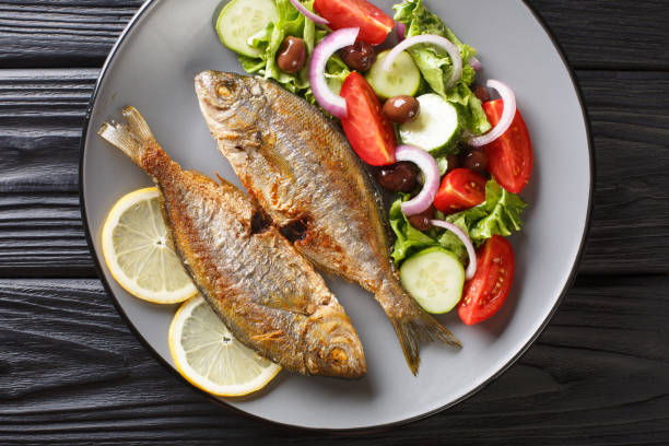 fried Salema porgy sea fish with fresh vegetable salad close-up on a plate on the table. Horizontal top view fried Salema porgy sea fish with fresh vegetable salad close-up on a plate. horizontal top view from above salpa stock pictures, royalty-free photos & images
