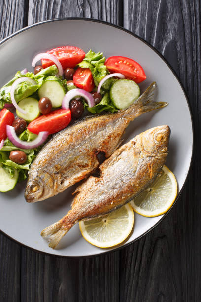 fried Salema porgy sea fish with fresh vegetable salad close-up on a plate on the table. Vertical top view fried Salema porgy sea fish with fresh vegetable salad close-up on a plate. Vertical top view from above salpa stock pictures, royalty-free photos & images