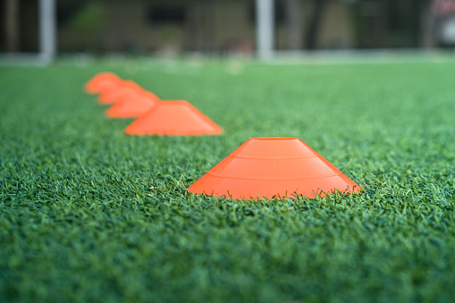 Football training cone is aligned on artificial grass pitch, ready for dribbling and movement training. Close up and selected focus photo at the object. Football time concept.
