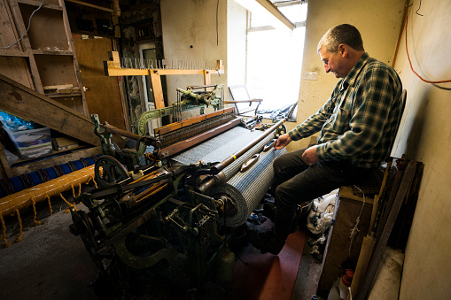 A male crofter busy working on his foot powered Hattersley loom at home weaves wool sheared, dyed and spun on the island according to Parliment bylaws to create distinctive tweed cloth fabric on the Isle of Lewis, Outer Hebrides, Scotland, UK