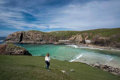 A female tourist wearing wellies, rubber boots, jacket and jeans walks along the cliffs overlooking Dalbeg Beach, Dail Beag, a hidden gem in a picturesque bay on the Atlantic Ocean on the northwest side of the Isle of Lewis in the Outer Hebrides, Scotland, UK, Europe