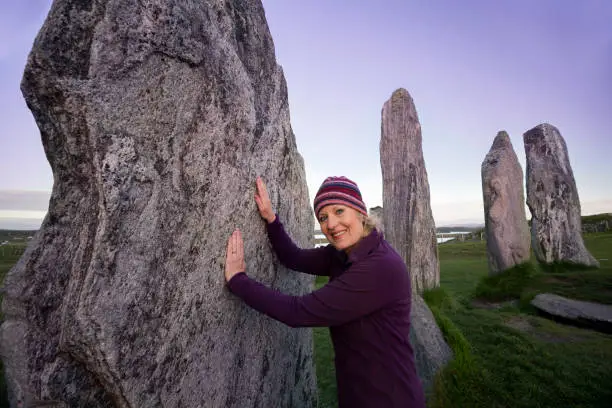 Female tourist in a striped woven hat and purple fleece jacket touches the stones at the famous Callanish Standing Stones, Outer Hebrides, Scotland, UK, Europe