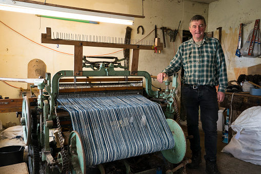 A male crofter stands beside his foot-powered Hattersley loom passed down through the generations at home used for weaving wool sheared, dyed and spun on the island according to Parliment bylaws to create distinctive tweed cloth fabric on the Isle of Lewis, Outer Hebrides, Scotland, UK