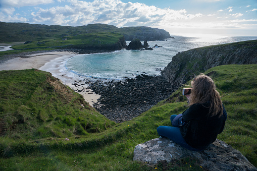 A female tourist wearing wellies, rubber boots, sits overlooking Dalbeg Beach, Dail Beag, a hidden gem in a picturesque bay on the Atlantic Ocean on the northwest side of the Isle of Lewis in the Outer Hebrides, Scotland, UK, Europe