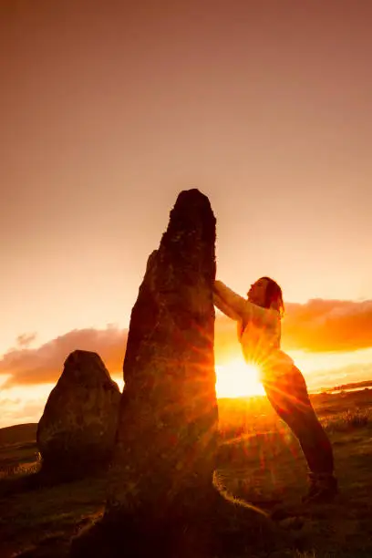 Tourist silhouetted against an orange sunset touches one of the rock formations at Callanish stones III, Calanais Standing Stones, Outer Hebrides, Isle of Lewis, Scotland, UK.