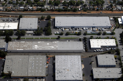 Aerial view of buildings In a manufacturing district. Many large warehouses with white and gray tattered roofs are visible in the foreground. Shipping trucks and Semi trucks are parking in loading bays. Employee cars are parking in a parking lot. Residential houses can be seen in the background, and trees are visible among the residences.