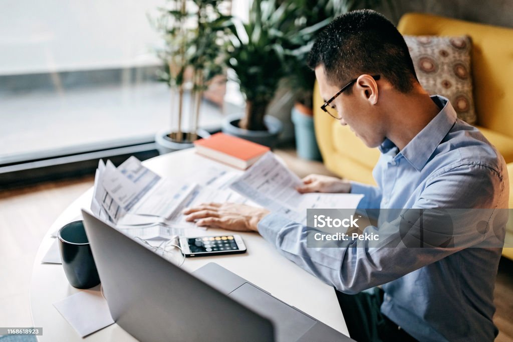 Japanese man calculating financial bills at home Japanese man with eyeglasses siting on floor in the living room and using smart phone and laptop for managing home finances Tax Form Stock Photo