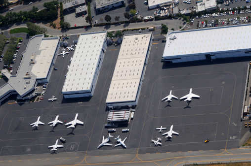 An aerial photo of an airport tarmac with a large cargo airplane parked at a gate. The airplane is surrounded by various airport vehicles and cargo containers. The tarmac is marked with yellow lines and has several other airplanes parked in the background. The background also includes a large parking lot and several buildings.