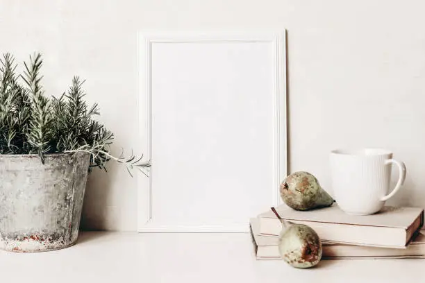 Photo of White vertical blank wooden frame mockup. Composition of rosemary herb in old metal flower pot, books, cup of coffee and pears on white table. Home decor. Rustic summer poster product design.