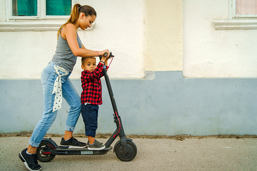 Young woman standing by the electric kick scooter board on the pavement in front of the wall of some house in the town riding driving her little small son or brother boy