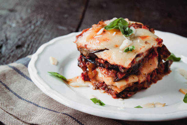 parmigiana di melanzane: melanzane al forno - italia, sicilia cousine. melanzane al forno con formaggio, pomodori e spezie su un piatto bianco. un piatto di melanzane è su un tavolo di legno - parmigiano foto e immagini stock