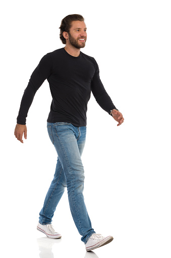Smiling young man in jeans, sneakers and black jersey is walking and looking away. Side view. Full length studio shot isolated on white.