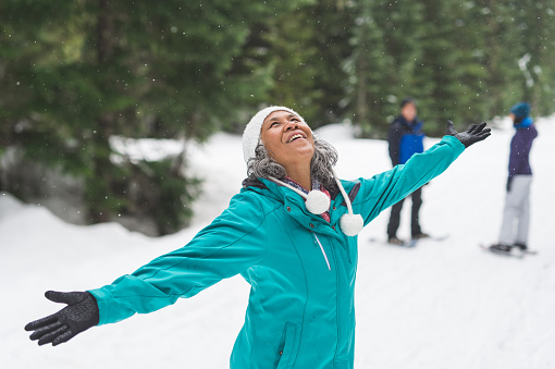 Beautiful ethnic woman in her 50s heads out for a snowshoeing adventure in the white forest with her husband and daughter, who are in the background. She is holding her arms in the air and looking up in wonderment.