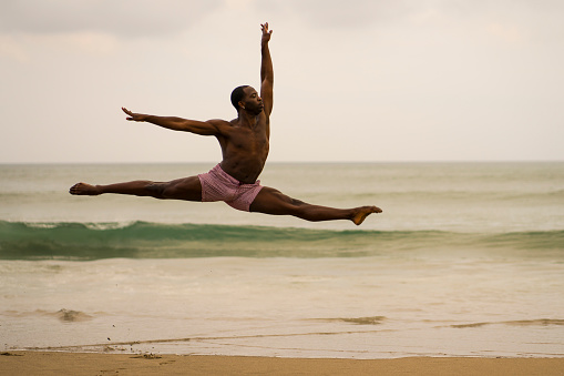 dramatic contemporary dance choreographer doing ballet beach workout . young attractive and athletic black African American man dancing on sunrise jumping elegant and with amazing plasticity