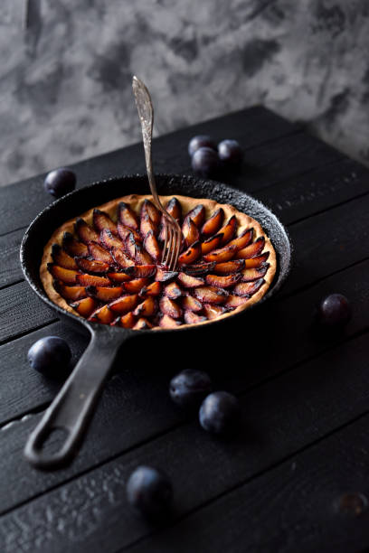 Baking failure. Burnt homemade plum pie in cast iron pan with fork in center on black background. Low key still life with natural lighting