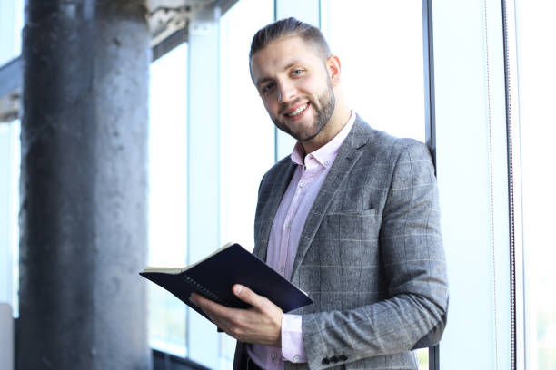 istock Good looking business man in casual clothes holding his note pad and looking at camera while standing in the office. 1168746719