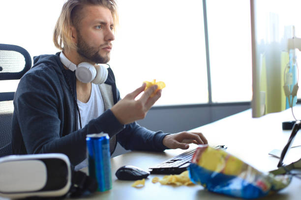 istock Concentrated young man in casual clothing using computer, streaming playthrough or walkthrough video and eating chips. 1168746875