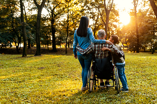 Photo of a man having happiness and smiling with his daughter and grandson on wheelchair at outdoor park. Mature man is happy with his family, child boy having fun talk, laughing together in nature. The veteran in a wheelchair came back from the army. Wife and son are glad to see him. A man in a wheelchair with his family.