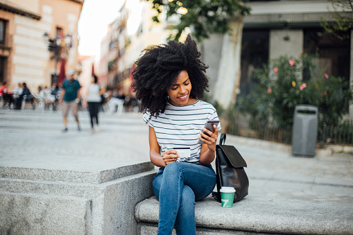 Beautiful, young African - American woman sitting on the street and shopping online.