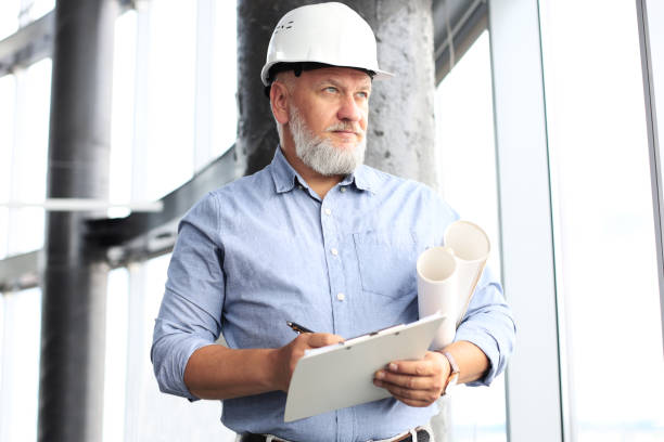 istock Confident mature business man in hardhat holding blueprint and looking away while standing indoors. 1168752217