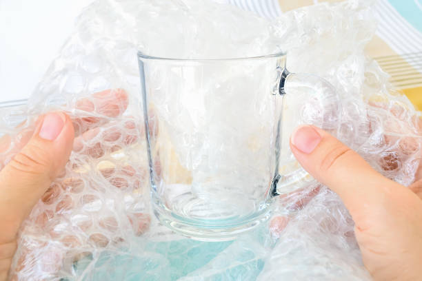 Woman hands packaging classic glass tea cup with transparent bubble pack on a white blue checkered tablecloth. Material for packing fragile items for safe transportation. Close-up.