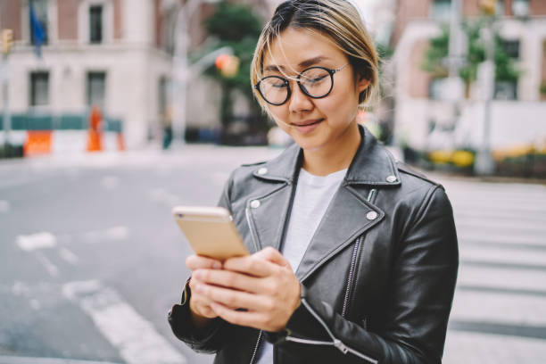 istock Young asian girl in eyewear texting feedback to friend chatting online while spending time outdoors in city, blonde chinese woman reading information about cafes from mobile strolling in downtown 1168760067