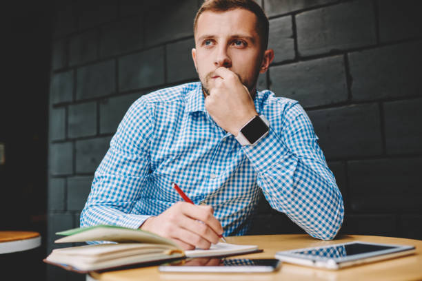 istock Pondering young man with modern smartwatch thinking on creative ideas for writing publication sitting in coffee shop.Thoughtful hipster student looking away while doing homework in cafeteria 1168762685