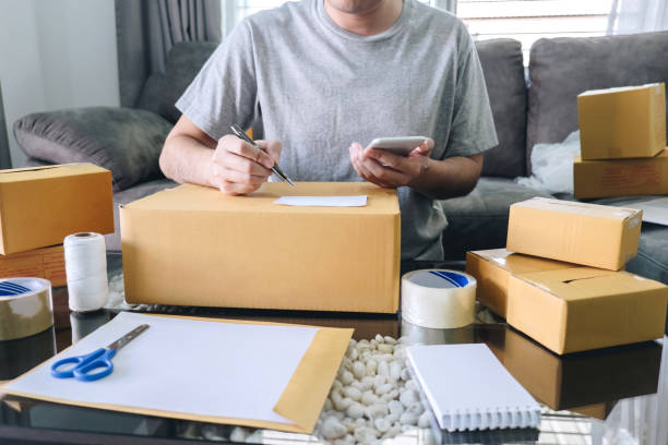 istock Young entrepreneur SME freelance man using smartphone receive order client and take note working with packaging sort box delivery online market on purchase order and preparing package product 1168773209