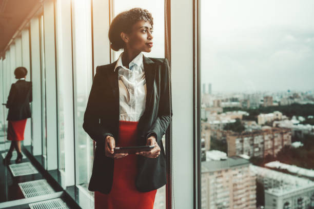 A businesswoman with digital tablet An African-American woman entrepreneur a digital tablet is leaning against a panoramic window of an office business skyscraper and looking in a distance; biracial businesswoman with a tablet pc bossy photos stock pictures, royalty-free photos & images