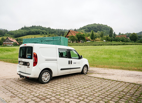 Ottenhofen Im Schwarzwald, Germany - Jul 28, 2019: Side view of Fiat Doblo electric van with SWEG logotype parked on a empty parking with forest and houses in background