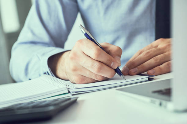 istock Businessman hand writing note on a notebook. Business man working at office desk. Close up of empty notebook on a blackboard with office supplies. 1168781720