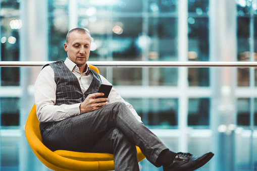 A confident adult man entrepreneur is sitting on an orange soft armchair in a modern office settings and using his smartphone; a caucasian businessman with the cellphone indoors, defocused background