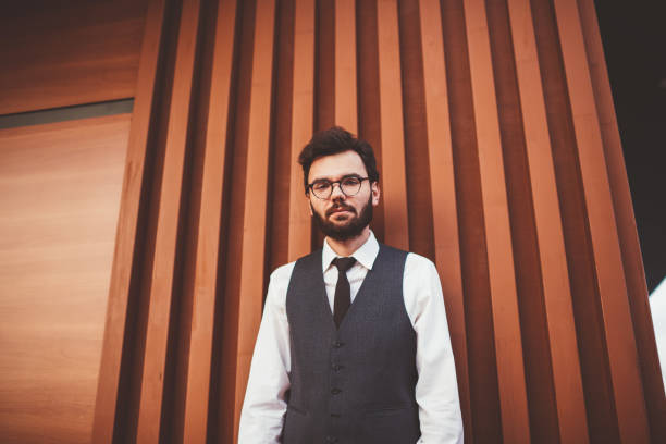 istock Caucasian male student in elegant formal apparel looking at camera while standing near wall on city street, young hipster guy in casual waistcoat spending free time on publicity area outdoors 1168785315