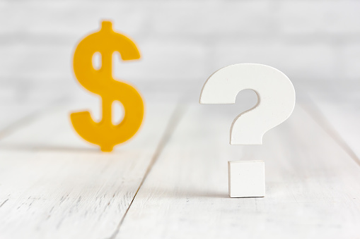 Question mark with dollar sign on white wood table over white brick background with copy space.