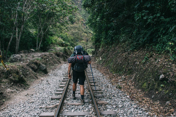 Back view of experienced young man traveler with backpack walking on railroad in forest during trip in South America.Wanderlust of male tourist with rucksack and trekking sticks for hiking Back view of experienced young man traveler with backpack walking on railroad in forest during trip in South America.Wanderlust of male tourist with rucksack and trekking sticks for hiking Sallqantay stock pictures, royalty-free photos & images