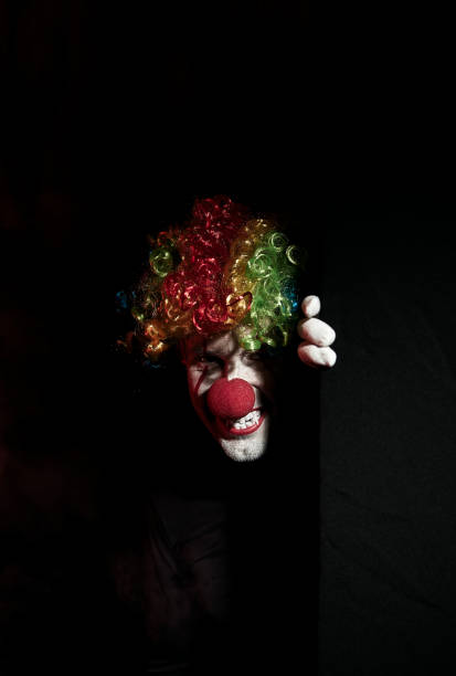 istock Scary clown peeping around the corner of a black wall. He is wearing a colored wig and sharp fangs. 1168800592