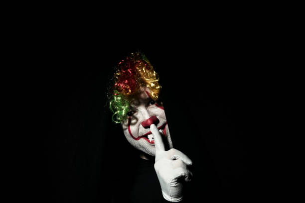 istock A terrible scary clown in a colored wig peeps out from behind black curtains. Shows a gesture of silence with a finger at the lips. 1168800622