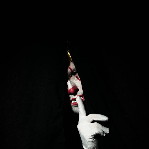 istock A terrible scary clown in a colored wig peeps out from behind black curtains. Shows a gesture of silence with a finger at the lips. 1168800623