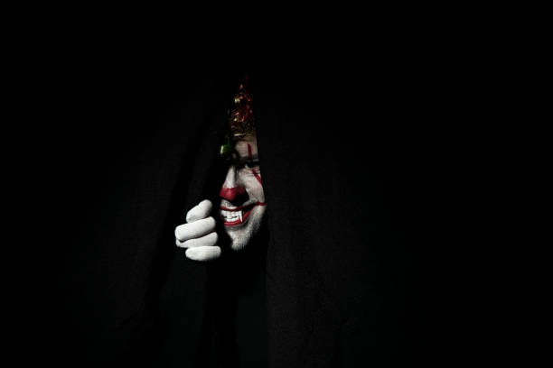 A terrible scary clown in a colored wig peeps out from behind black curtains.