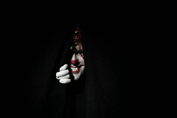 A terrible scary clown in a colored wig peeps out from behind black curtains. stock photo