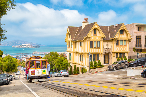 Historic Powell-Hyde cable car climbing up steep hill in central San Francisco with famous Alcatraz Island in the background on a sunny day with blue sky, USA.
