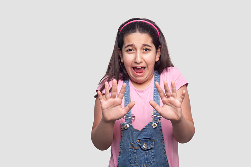 Scared sad brunette young girl in casual style, pink t-shirt and blue denim overalls standing with blocking hand and afraid face looking at camera. indoor studio shot isolated on light gray background