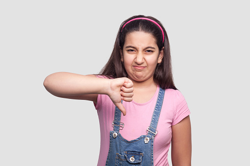 Dislike. Portrait of displeased sad brunette young girl in casual pink t-shirt and blue denim overalls standing and looking at camera with thumbs down. indoor studio shot, isolated on gray background.