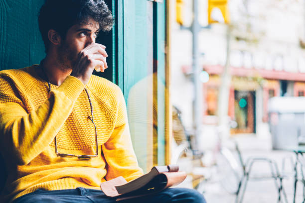 istock Bearded creative young man drinking tasty coffee while looking out of window and thinking on new ideas for creating schedule.Thoughtful male dressed in casual yellow sweater enjoying beverage 1168817182