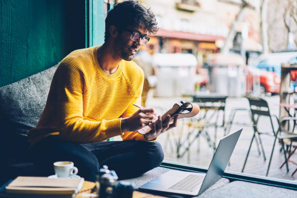 smart e-learning in optischen brillen, die online-webinar beobachten und notizen im notizblock während des e-lernens im coworking space machen. cleverer junger mann schreibt text in notizbuch von internet-website - netbook stock-fotos und bilder