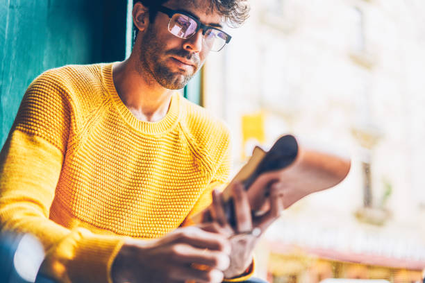 istock Cropped image of smart young man in optical eyegasses reading information from notepad.Pensive hipster guy dressed in yellow sweater learning text from notebook sitting in coworking space 1168817206