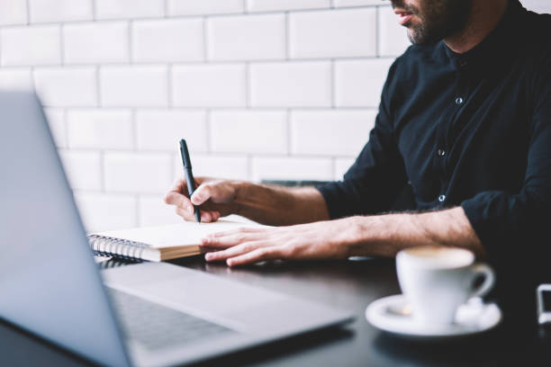 istock Cropped image of stylish businessman dressed in black shirt making notes in notepad working at modern laptop computer in coffee shop interior.Student writing in notebook sitting at netbook device 1168817252