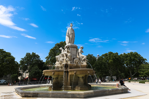 Fontaine Pradier in Esplanade Charles-de-Gaulle in the french city Nime in Frances. Unidentified people sit on the square near the fountain in the summer on a sunny day against a blue sky and park