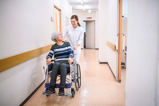 Young female Japanese nurse talking and laughing with senior woman as she pushes her wheelchair down hospital corridor.